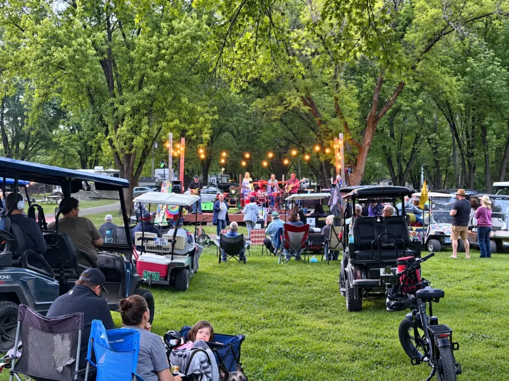 Campers watching a live band beneath string lights and shade trees at Holiday Acres